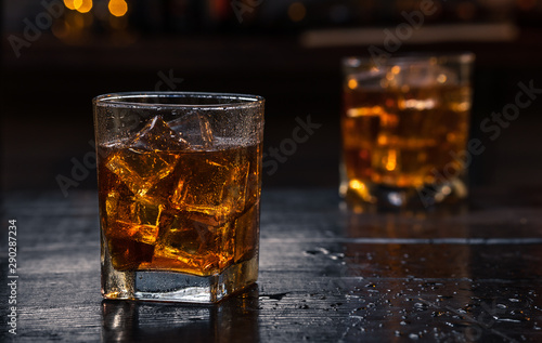 Two Glasses of Whisky (whiskey), bourbon, ice tea or coke (cola) soda with ice and water drops on wood table at dark background with copy space, close up