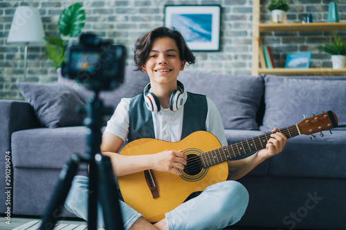 Portrait of teenage blogger recording video at home holding guitar smling using professional camera on tripod. Modern technology and vlogging concept. photo