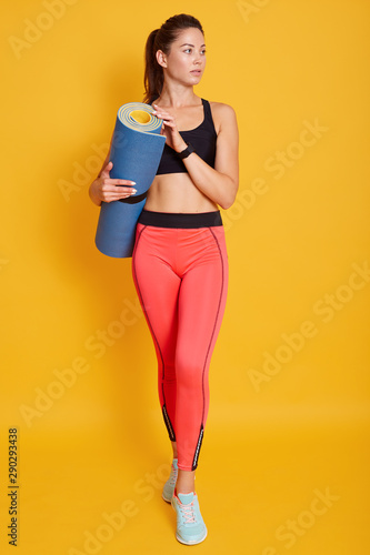 Full length portrait of beautiful athletic young woman holding yoga mat in hands, looking aside, ready for working out, sporty female posing isolated over yellow background. Fitness concept. photo