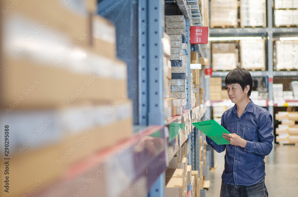 Asian manager man doing stocktaking of products management in cardboard box on shelves in warehouse using clipboard file and pen. Male professional assistant checking stock in factory. 