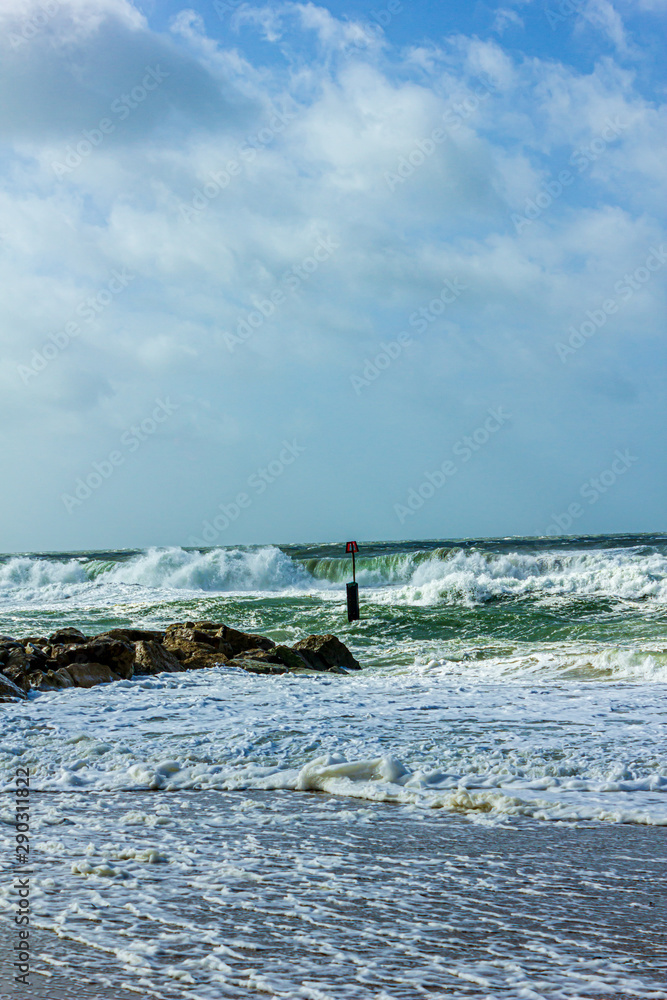 Fototapeta premium Crashing waves on a stony groyne (breakwater) during a massive storm under a majestic blue sky and white clouds