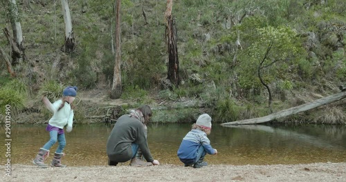 a mother is teaching her young children how to skip stones across a river. They are squatting on the river bank while the woman throws a stone, the kids throw stones too photo