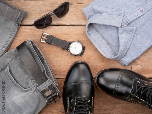 Creative fashion design outfits for men casual clothing set on wooden background include black boots, gray shirt, watch, sunglasses, jeans and belt leather. Top view
