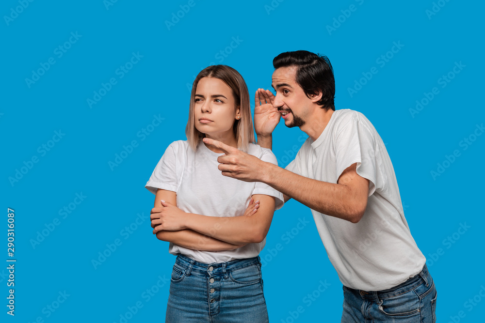 Bearded man with mustaches in white t-shirt tells secret to a young blond girl in white t-shirts and blue jeans standing isolated over blue background. Concept of secret telling. Bearded man with mustaches in white t-shirt tells secret to a young blond girl in white t-shirts and blue jeans standing isolated over blue background. Concept of secret telling.