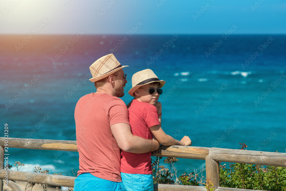 Dad and son in sun hats and sunglasses and similar clothes standing on cliff against wonderful sea view. They smiling and enjoying their summer vacations.