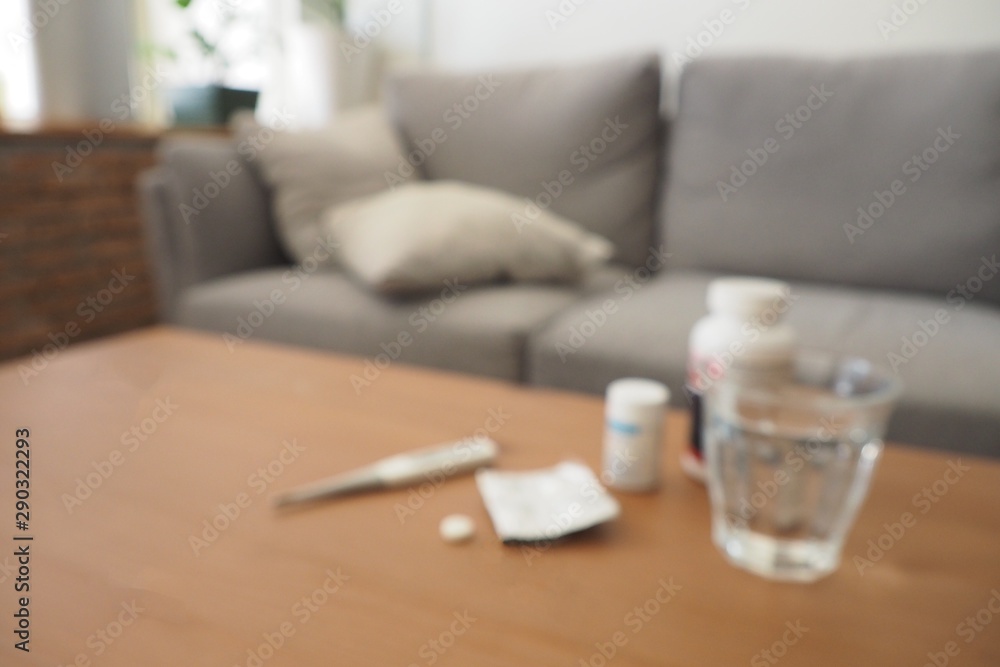 Blurred focus of glass of water, medical thermometer and bottles of pills on the table in the room. Taking pills and drugs concept when feeling sick and danger of overdose can destroy health.