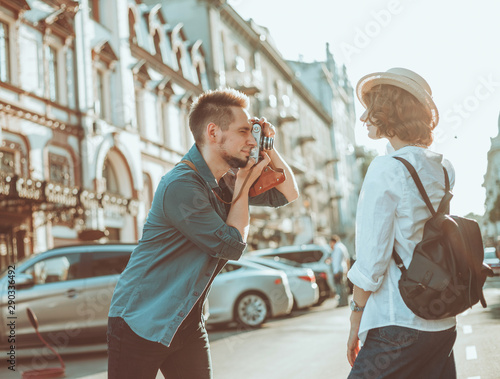 Young cheerful couple of hipsters are photographed on a retro camera while walking around the city. Male photographer photographs a stylishly dressed young woman