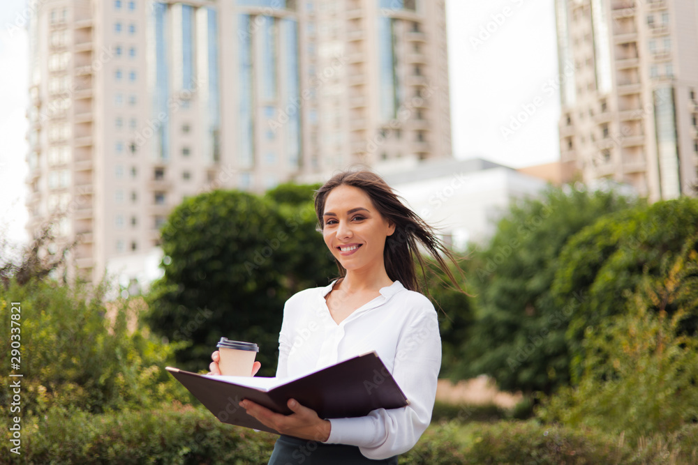 Businesswoman. Young attractive female manager wearing a skirt and a blouse holding folder with documents and coffee cup at background of the city landscape. All in business, coffee on the go Businesswoman. Young attractive female manager wearing a skirt and a blouse holding folder with documents and coffee cup at background of the city landscape. All in business, coffee on the go