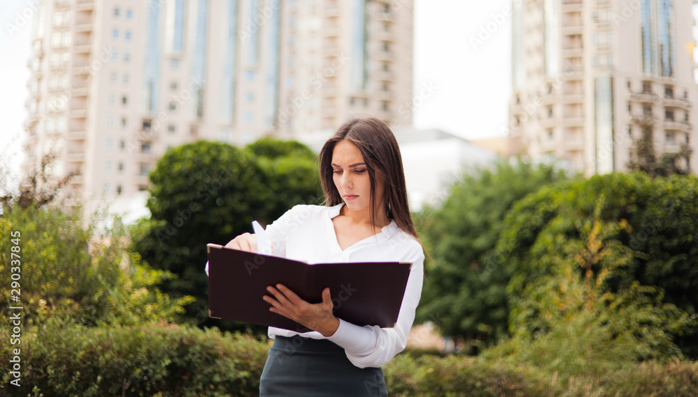 Fototapeta premium Businesswoman. Young attractive female manager wearing a skirt and a blouse holding folder with documents at background of the city landscape. All in business