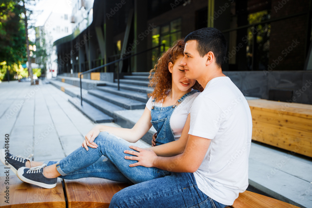 A young couple is sitting on a bench in a modern city, hugging and smiling. A young couple is sitting on a bench in a modern city, hugging and smiling.