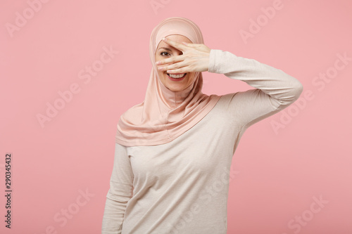 Cheerful young arabian muslim girl in hijab light clothes posing isolated on pink background. People religious Islam lifestyle concept. Mock up copy space. Hiding, peeping, covering eyes with hand.