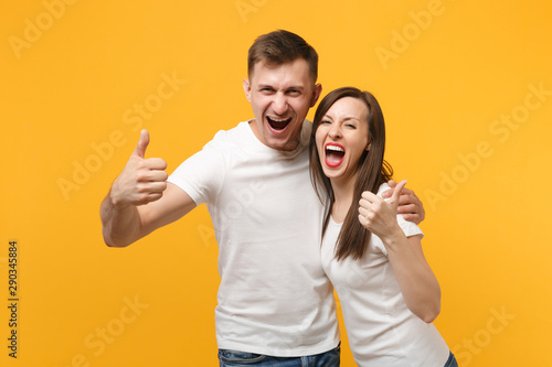 Joyful young couple two friends guy girl in white empty blank design t-shirts posing isolated on yellow orange background. People lifestyle concept. Mock up copy space. Showing thumbs up, screaming.