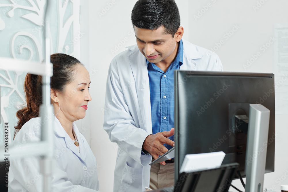 Indian male doctor showing something on tablet pc to his Asian colleague while she sitting at the table at office Indian male doctor showing something on tablet pc to his Asian colleague while she sitting at the table at office