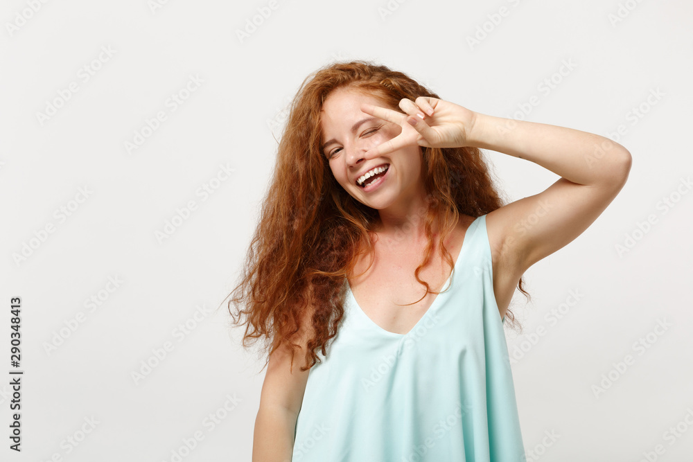 Young cheerful redhead woman in casual light clothes posing isolated on white wall background in studio. People sincere emotions lifestyle concept. Mock up copy space. Showing victory sign, blinking.