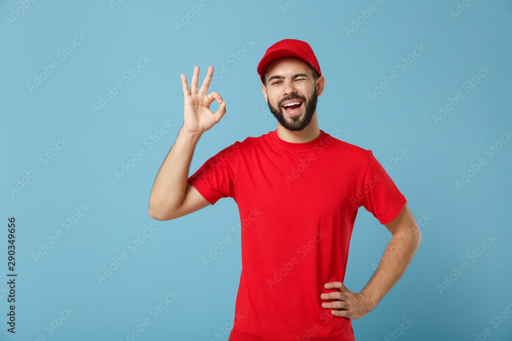 Delivery man in red uniform workwear isolated on blue wall background, studio portrait. Professional male employee in cap t-shirt print working as courier dealer. Service concept. Mock up copy space. Delivery man in red uniform workwear isolated on blue wall background, studio portrait. Professional male employee in cap t-shirt print working as courier dealer. Service concept. Mock up copy space.