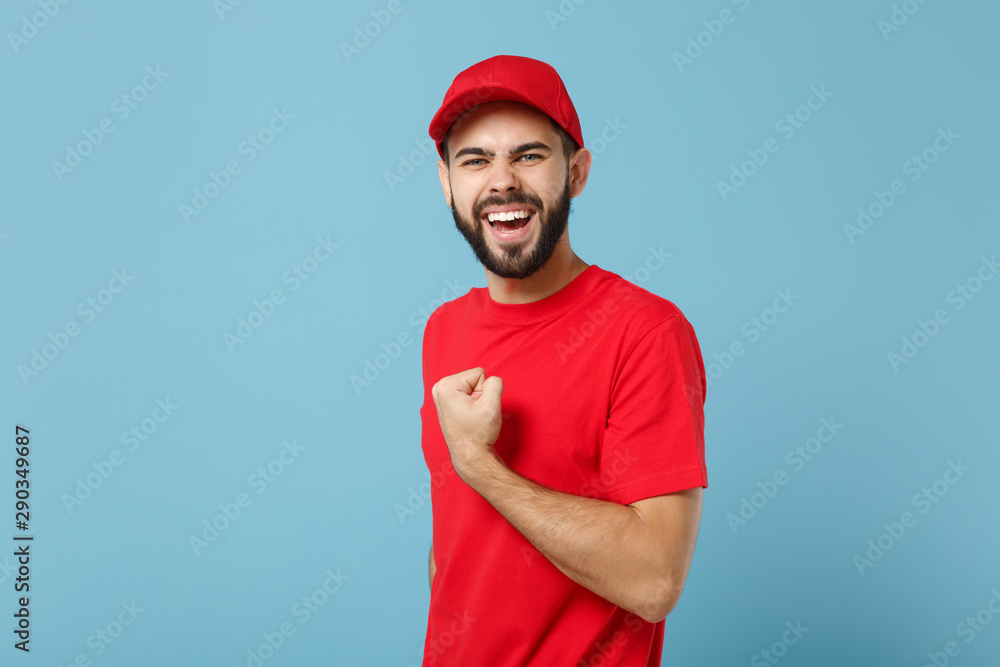 Delivery man in red uniform workwear isolated on blue wall background, studio portrait. Professional male employee in cap t-shirt print working as courier dealer. Service concept. Mock up copy space. Delivery man in red uniform workwear isolated on blue wall background, studio portrait. Professional male employee in cap t-shirt print working as courier dealer. Service concept. Mock up copy space.