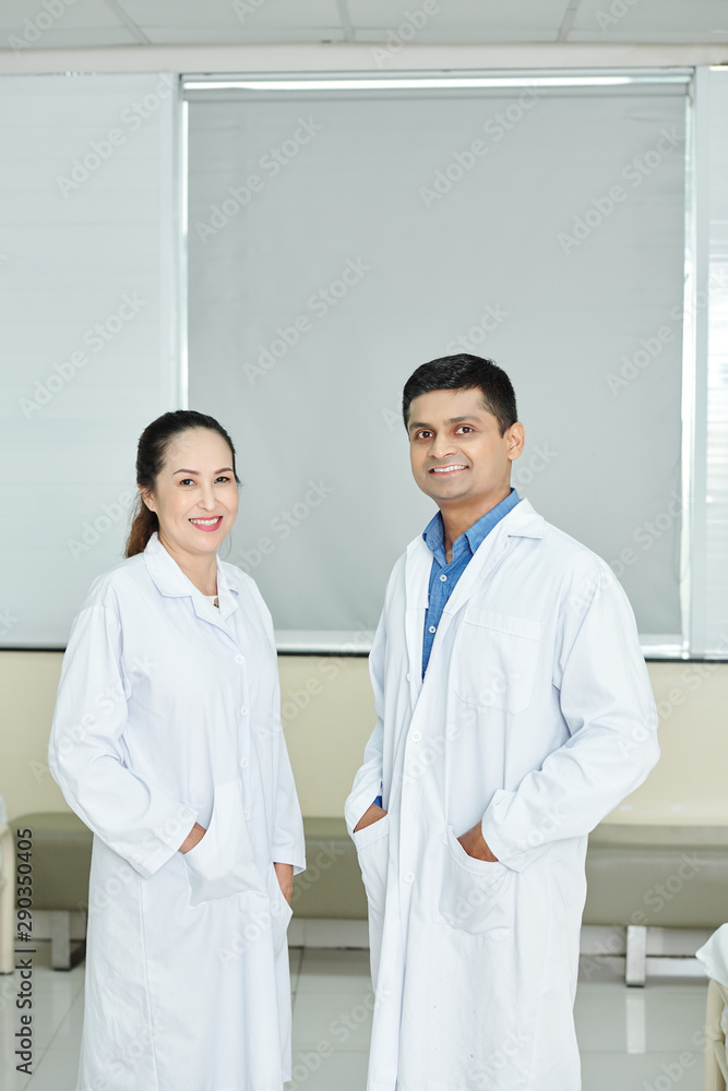 Portrait of Indian male doctor in white coat standing together with his Asian nurse and they smiling at camera at hospital Portrait of Indian male doctor in white coat standing together with his Asian nurse and they smiling at camera at hospital