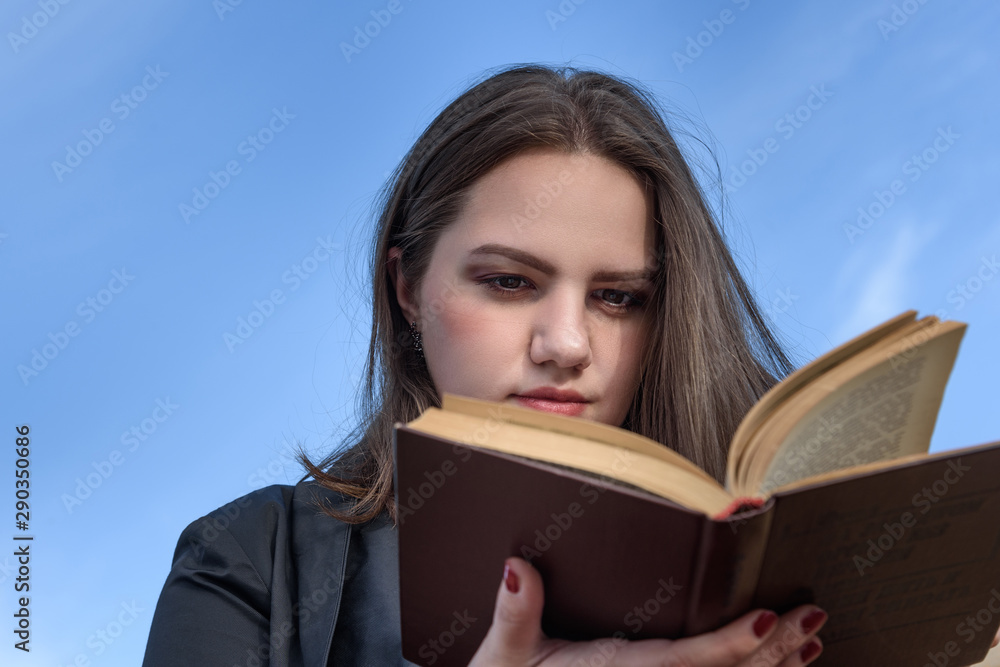 Brunette girl reading book on bench on the street. Girl in black jacket with long hair with book . Plus size model Brunette girl reading book on bench on the street. Girl in black jacket with long hair with book . Plus size model