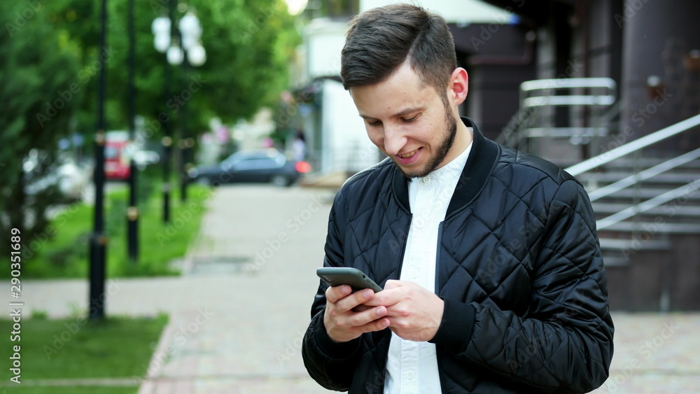Handsome young bearded man uses the phone on the street in, typing text, chatting with friends - obrazy, fototapety, plakaty 