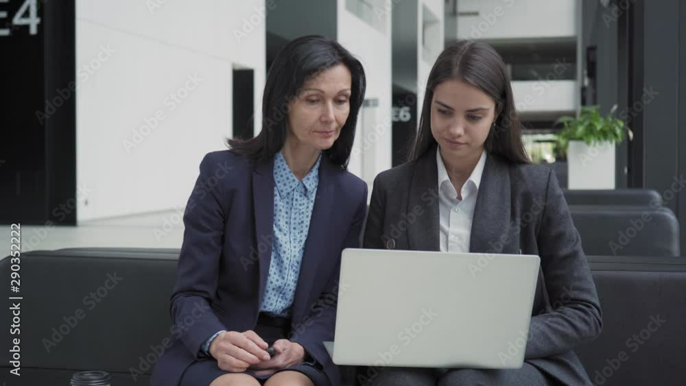 Medium shot of two businesswomen sitting on sofa in office hall and planning project together using laptop. Middle aged entrepreneur mentoring young female colleague Medium shot of two businesswomen sitting on sofa in office hall and planning project together using laptop. Middle aged entrepreneur mentoring young female colleague