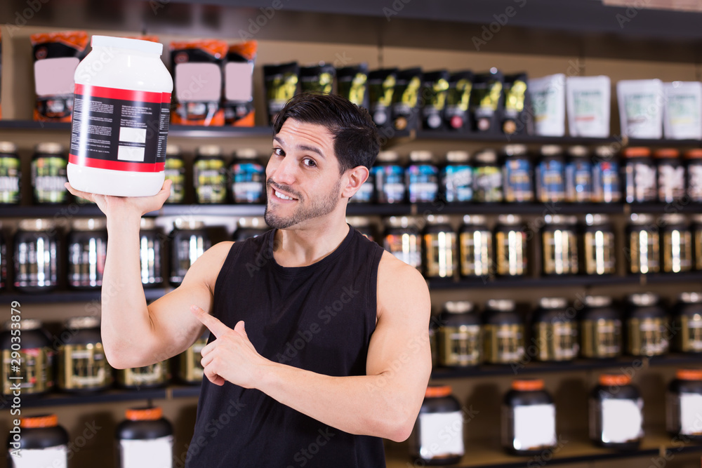 Positive muscular male seller holding big pot of sport nutrition
