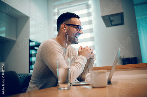 Side view of beautiful positive man dressed casual sitting at dining table in kitchen and having video call over laptop with his girlfriend. On table next to laptop are glass of water and coffee.