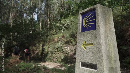 Way of St. James sign and pilgrims walking on a forest footpath. Yellow scallop sign pilgrimage to Santiago de Compostela