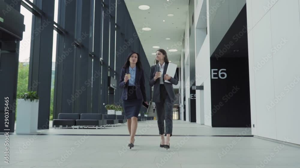 Full length slow motion shot of two businesswomen, young and middle aged, in formal suits walking down office hall together and talking. Colleagues holding takeaway coffee cups, cell phone and laptop Full length slow motion shot of two businesswomen, young and middle aged, in formal suits walking down office hall together and talking. Colleagues holding takeaway coffee cups, cell phone and laptop