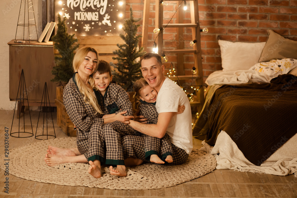 happy family, father, mother and two children are sitting on the floor in the room by the bed and hugging, people in pajamas are waiting for a merry Christmas