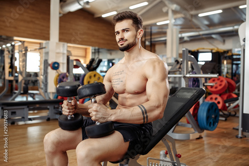 Muscular athletic young man bodybuilder fitness model focusing on exercise with dumbbell in the gym.