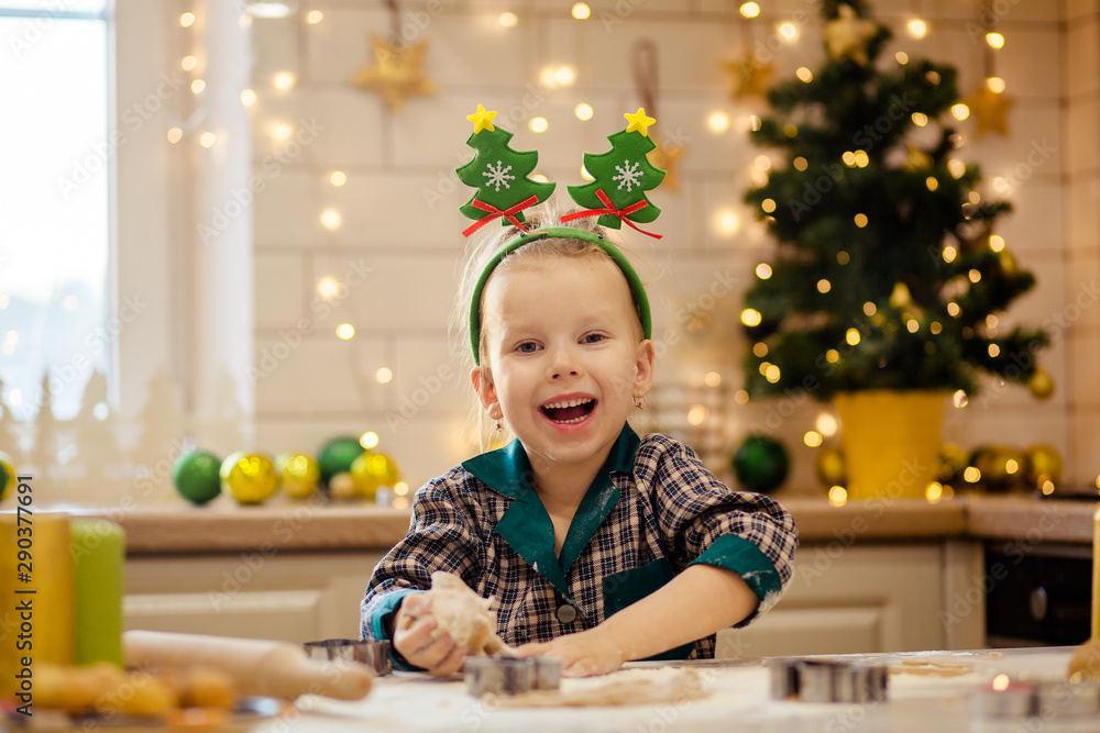 Kids bake Christmas gingerbread. Happy beautiful girl in pajamas preparing dough for new year cookies to celebrate Christmas. White kitchen with a Christmas tree and lights on a snowy winter day. Kids bake Christmas gingerbread. Happy beautiful girl in pajamas preparing dough for new year cookies to celebrate Christmas. White kitchen with a Christmas tree and lights on a snowy winter day.