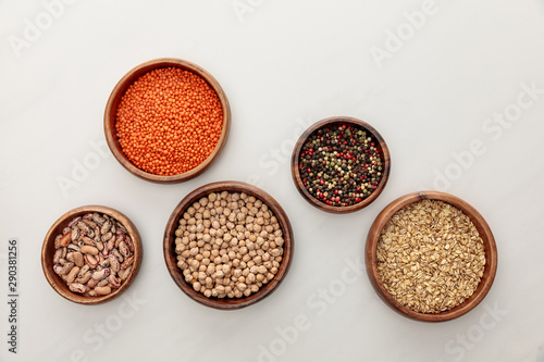 top view of wooden bowls with red lentil, beans, chickpea, oatmeal and peppercorns on white marble surface