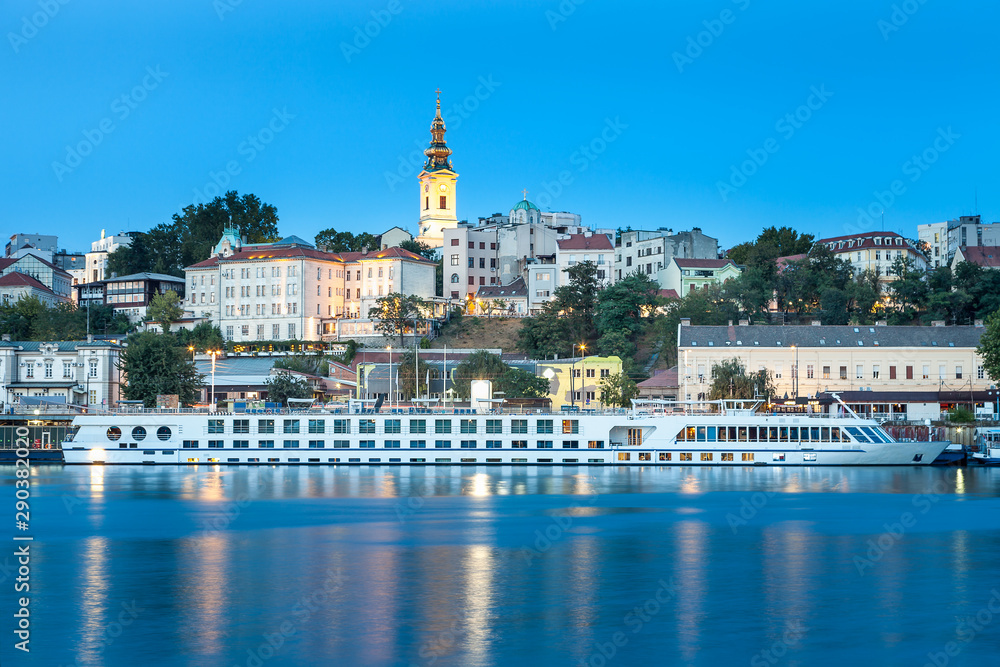 Blue hour view of Belgrade riverfront with Sava river in the foreground, holy archangel Michael cathedral at the back and impressive, large cruiser ship in the harbor