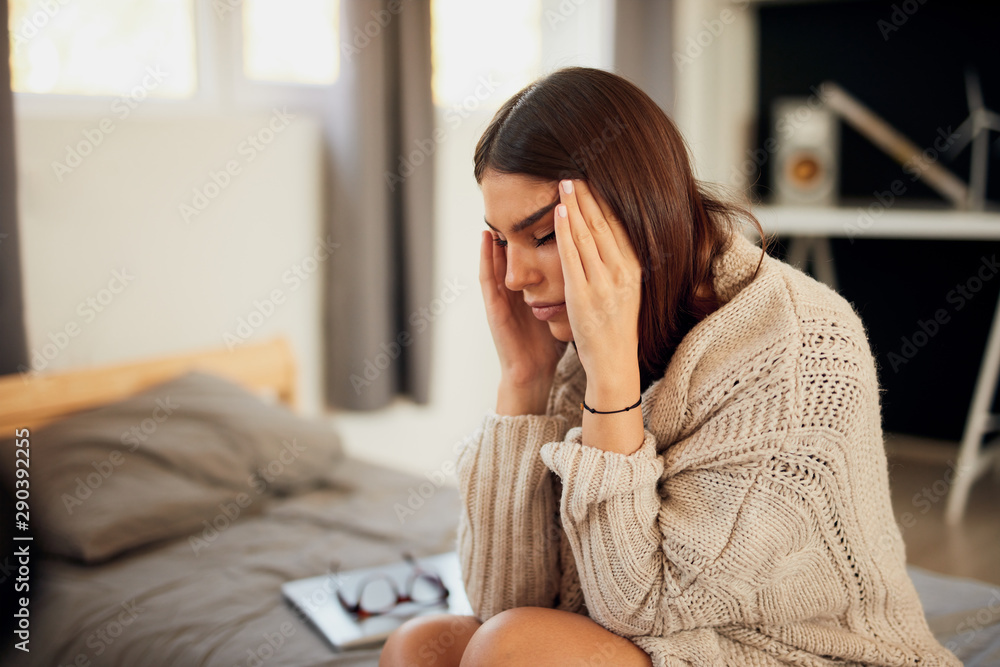 Attractive caucasian brunette dressed in beige sweater sitting on bed in bedroom and having headache.