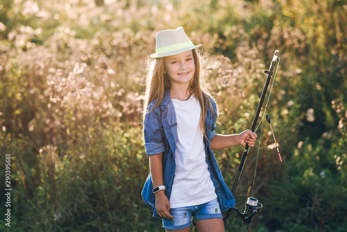 pretty little girl in shorts and hat on sunset background holding fishing rod in her hands