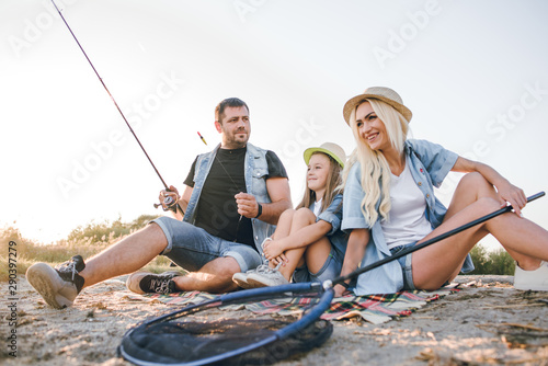 Happy young family fishing on the lake. They smile, hold fishing rods and fishnets in their hands. The little daughter helps her parents fish. Wonderful landscape of the lake at sunset