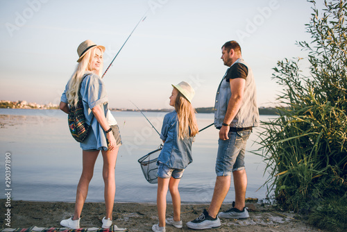 Happy young family fishing on the lake. They smile, hold fishing rods and fishnets in their hands. The little daughter helps her parents fish. Wonderful landscape of the lake at sunset