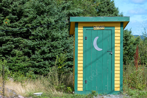 Yellow outhouse with green trim and green door. There's a white half moon painted on the exterior of the door.  A sign hangs over the door with the word outhouse. Green trees are in the background.