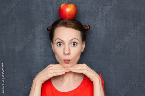 Fotografi Portrait of cute surprised woman with red apple on head