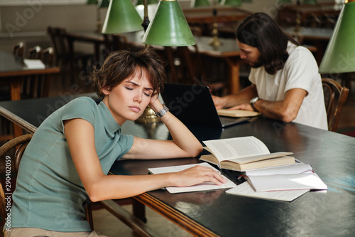 Young exhausted student tiredly sleeping on desk with books during study in library of university