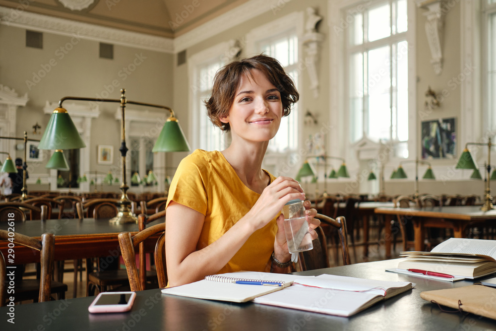 Young pretty smiling female student with water in hand joyfully looking in camera while study in library of university