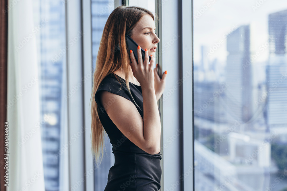 Elegant young female business owner holding documents talking on mobile phone discussing contract conditions standing in office. Elegant young female business owner holding documents talking on mobile phone discussing contract conditions standing in office.