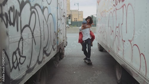 Mixed Race man carrying woman piggyback on skateboard near trucks
