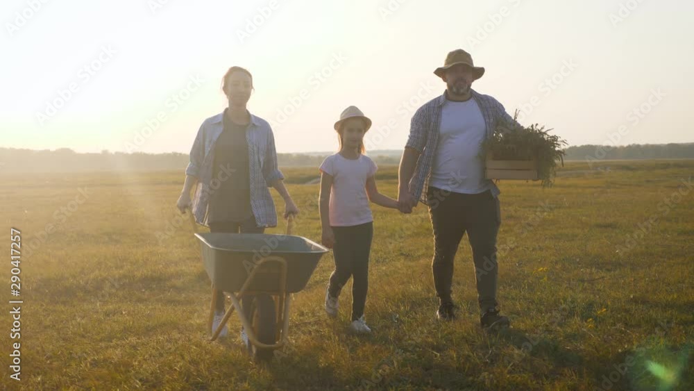 Young family having fun outdoors in their farm. Gardener woman pushing wheelbarrow with vegetables at sunset. Family working at the countryside. Silhouette workers go at sunset. Happy family concept. Young family having fun outdoors in their farm. Gardener woman pushing wheelbarrow with vegetables at sunset. Family working at the countryside. Silhouette workers go at sunset. Happy family concept.