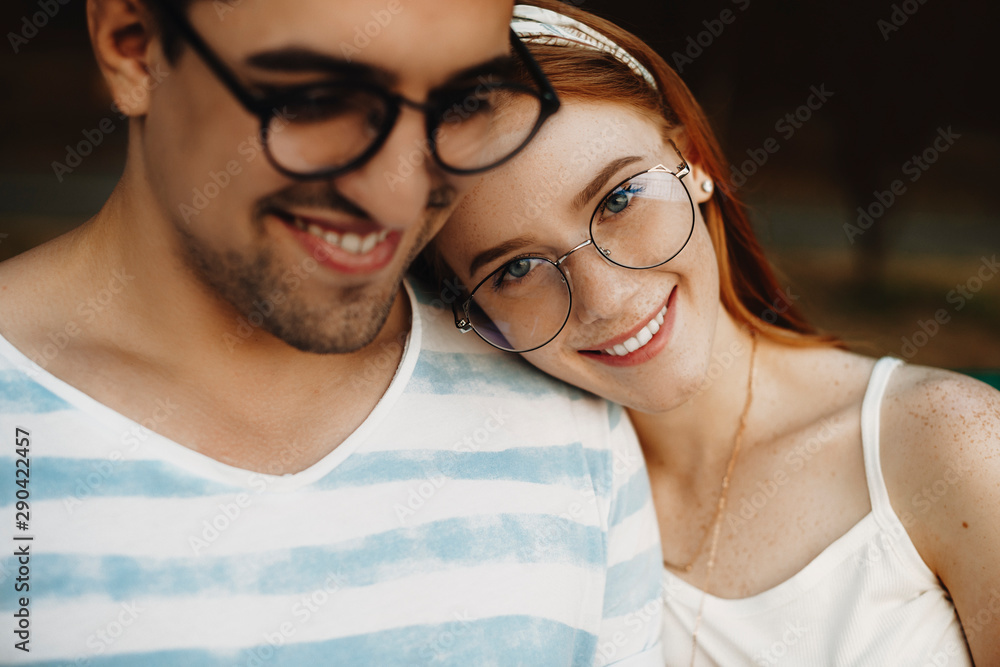 Obraz premium Close up portrait of a young couple sitting closely while smiling. Red haired girl with freckles looking at camera while leaning head on her boyfriend.