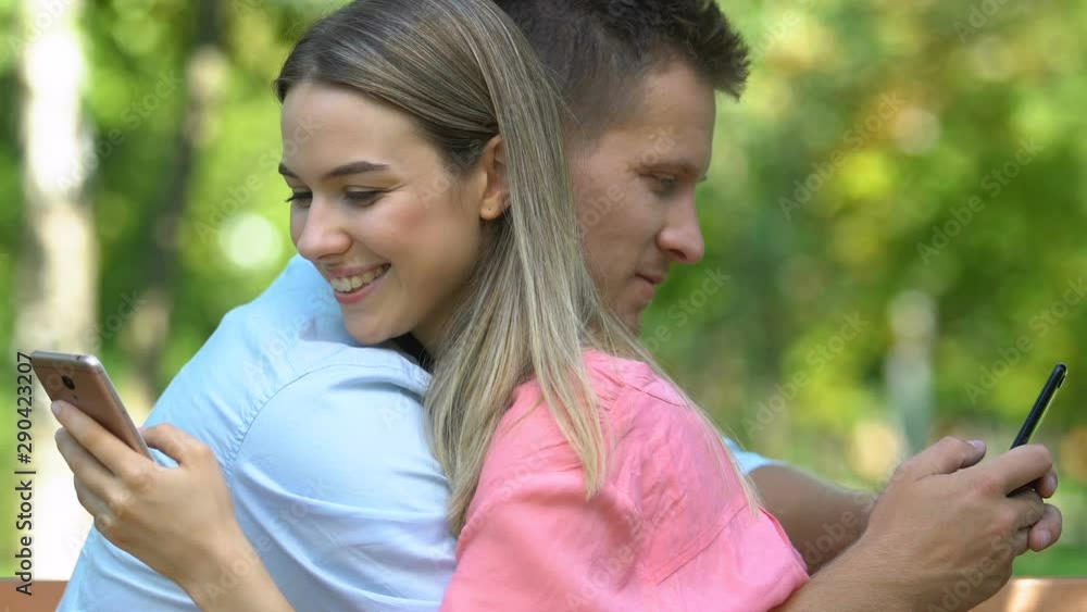 Couple tenderly hugging sitting on bench and chatting on smartphones, close-up Couple tenderly hugging sitting on bench and chatting on smartphones, close-up