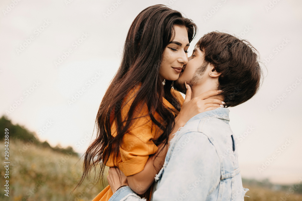 Close up of a lovely young couple embracing outdoor on a field. Charming young brunette embracing with closed eyes her boyfriend while he is kissing her. Close up of a lovely young couple embracing outdoor on a field. Charming young brunette embracing with closed eyes her boyfriend while he is kissing her.