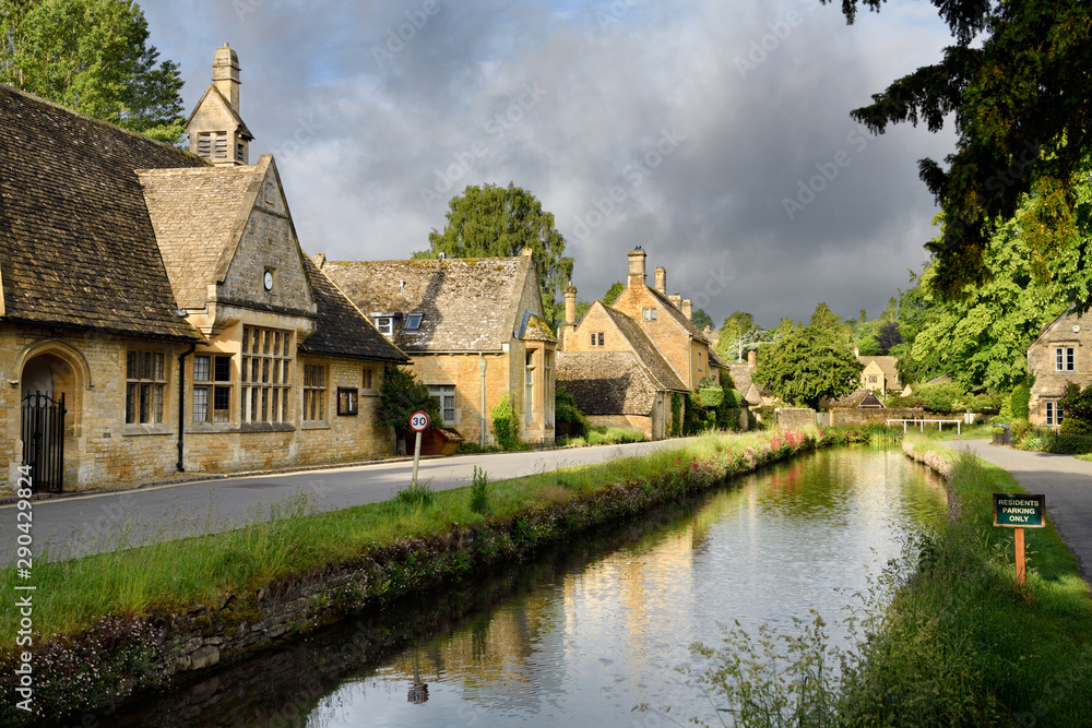 Morning sun on yellow Cotswold limestone buildings of Lower Slaughter on the River Eye with dark clouds Gloucestershire England