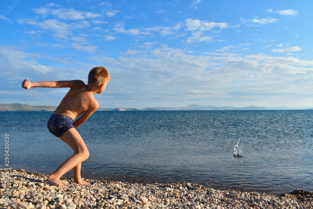 Boy in swimming trunks on  lake throws stones into  water. Teenager plays on seashore. Summer vacation with parents on lake on fresh air.