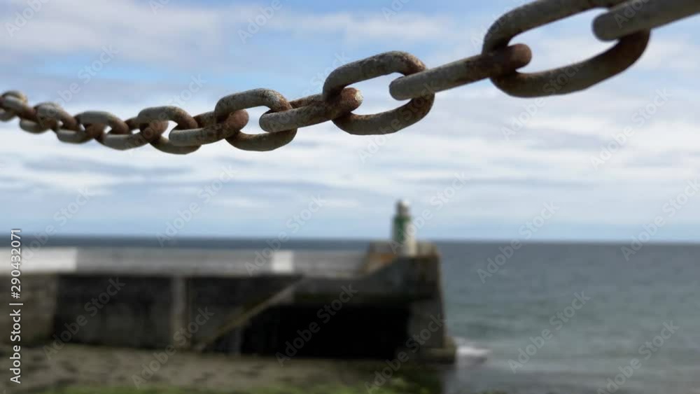 Chain Link In Focus With Waves Hitting Stone Pier With Small Lighthouse At Laxey, Isle Of Man. LOCKED OFF Chain Link In Focus With Waves Hitting Stone Pier With Small Lighthouse At Laxey, Isle Of Man. LOCKED OFF